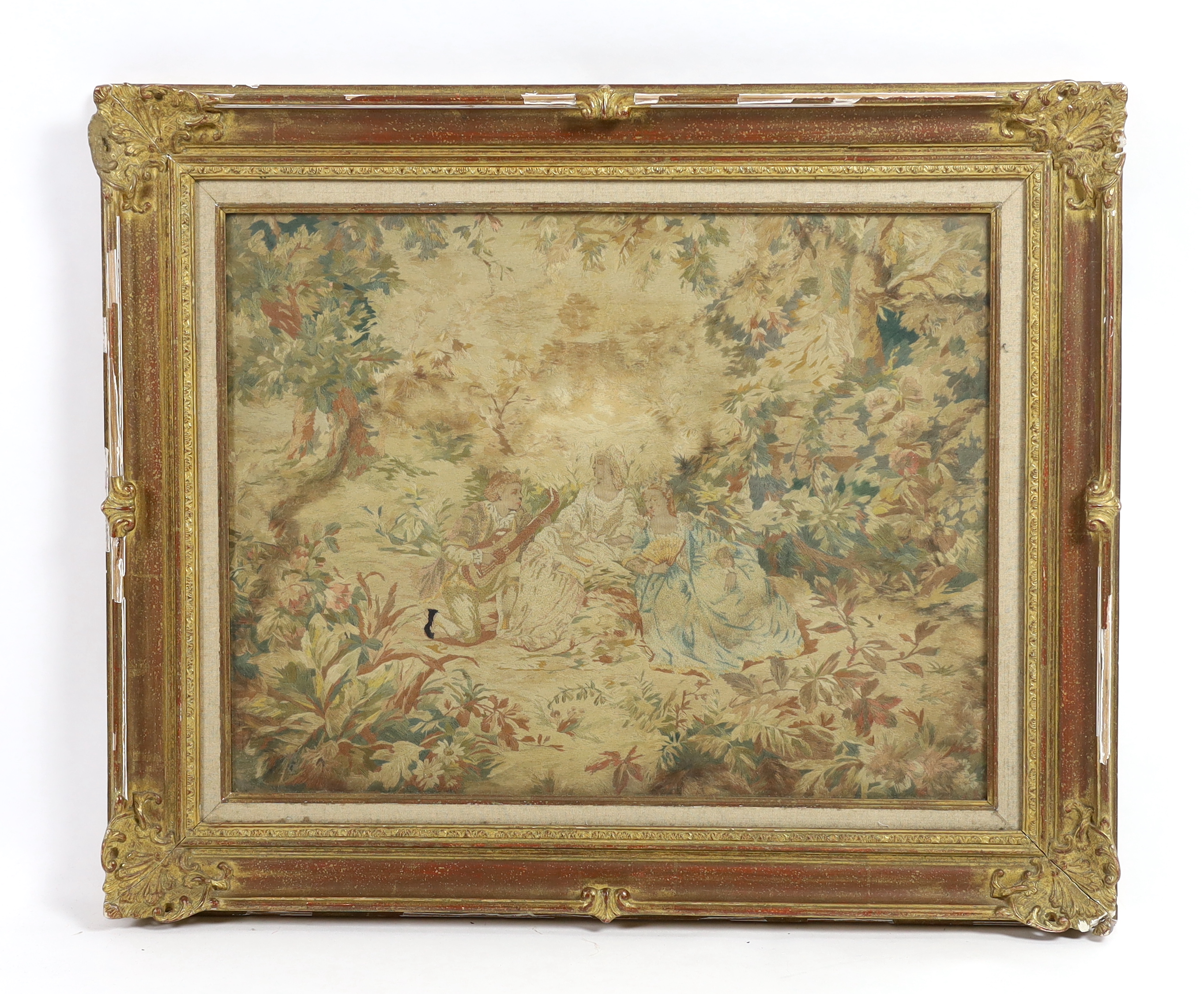 A mid 19th century framed ornate silk embroidery panel of two seated ladies being serenaded by a young musician in a luscious wooded landscape, 64cm wide, 49cm high